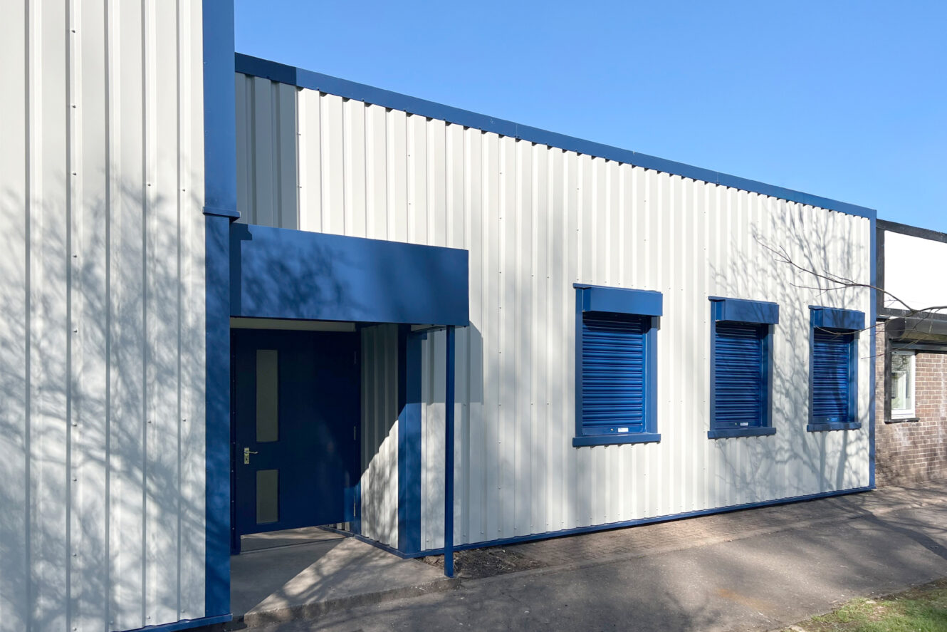 A white industrial building with blue trim, three blue-shuttered windows, a blue door, and corrugated metal walls under a clear sky.