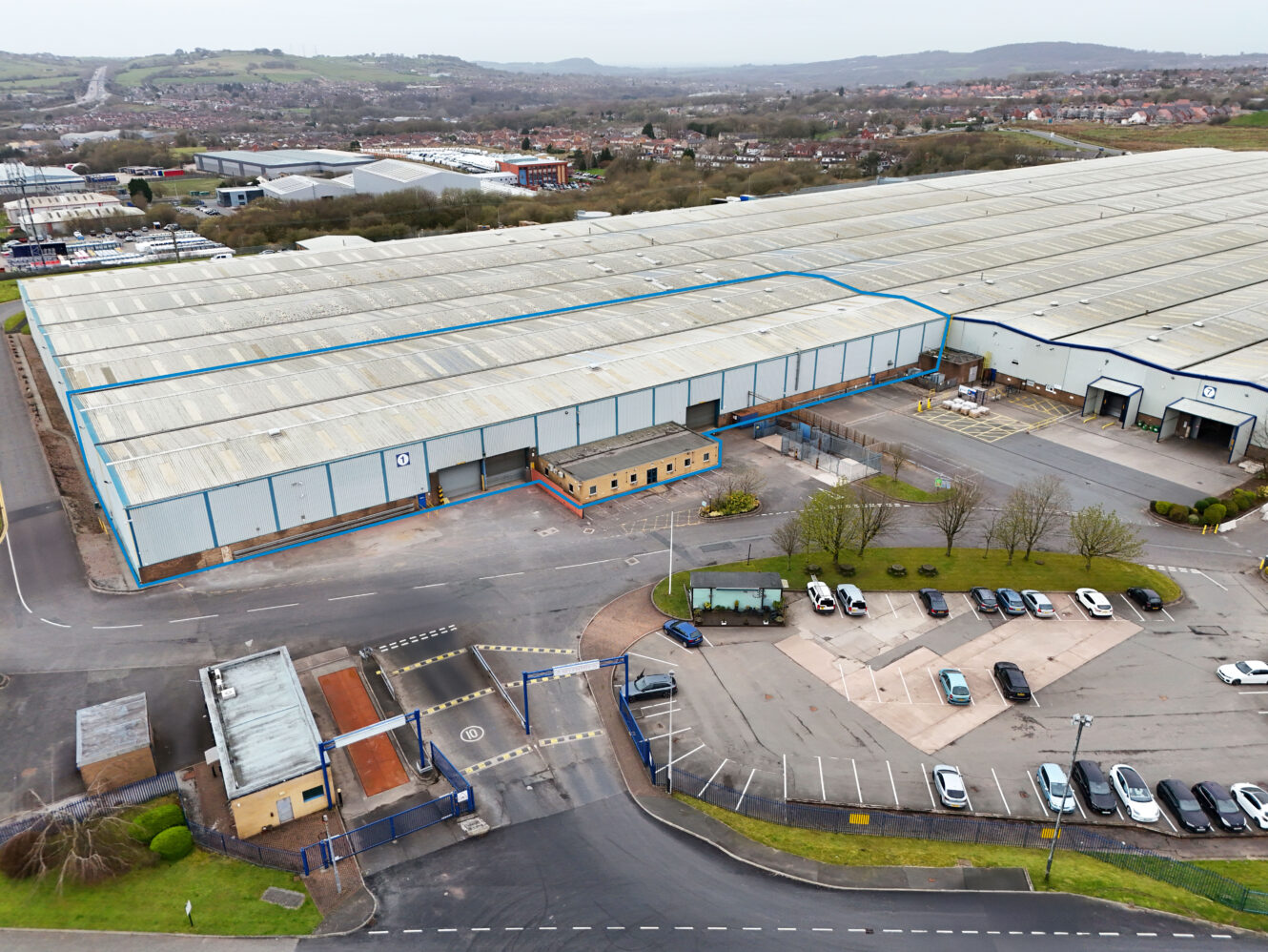 A large industrial warehouse complex with a parking lot in front, security gatehouse at the entrance, and surrounding roads and greenery.