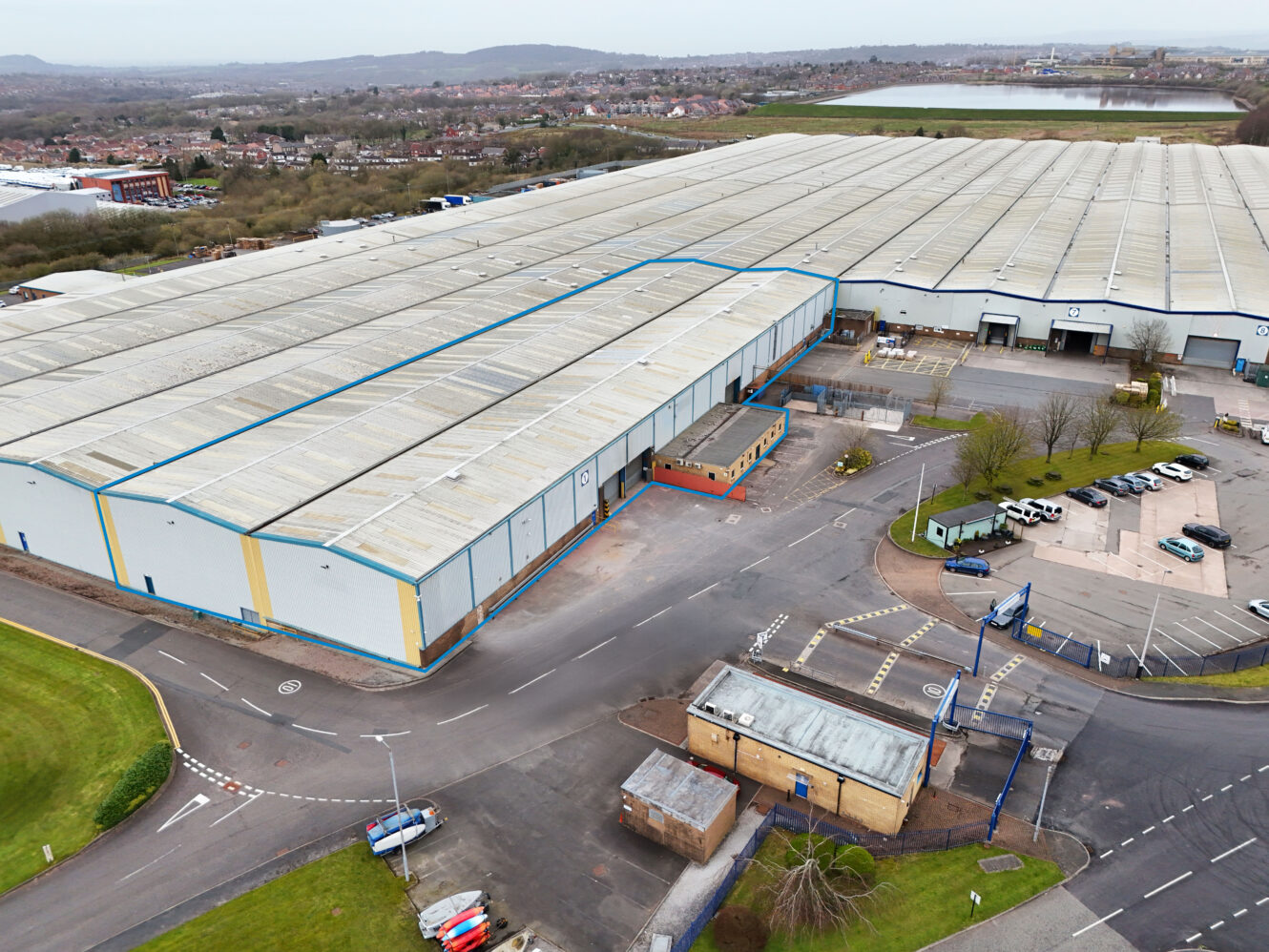 Aerial view of a large industrial warehouse with multiple loading bays, adjacent parking lot, surrounding roads, and nearby green areas.