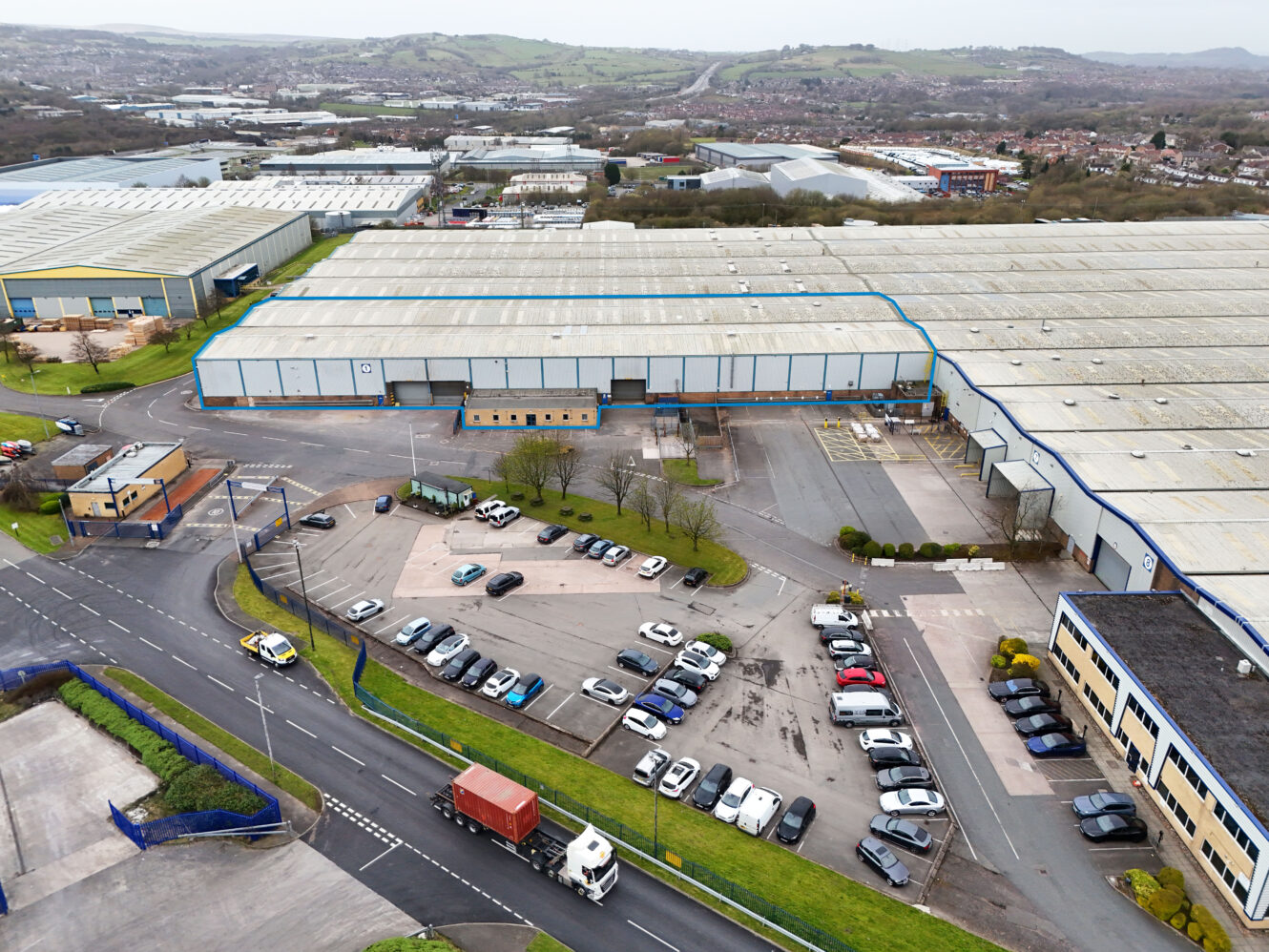 Aerial view of a large industrial warehouse complex with multiple buildings, parking lots with cars, trucks, and surrounding roads in a suburban area with hills in the background.