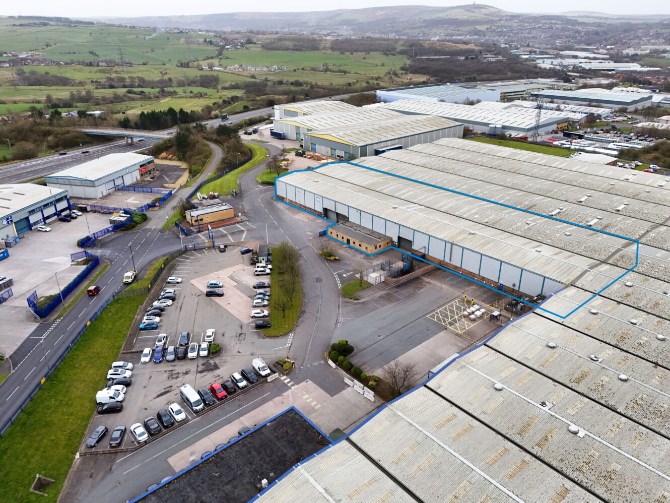 Aerial view of an industrial estate with several large warehouses, parking lots with cars, roads, and green hills in the background.