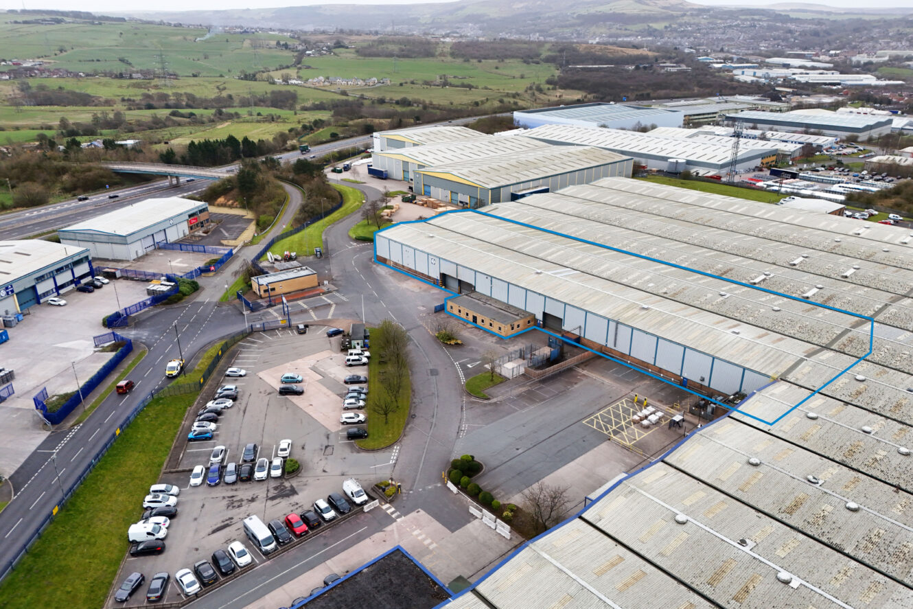 Aerial view of an industrial estate with several warehouses, parking lots filled with cars, roads, and green hills in the background.