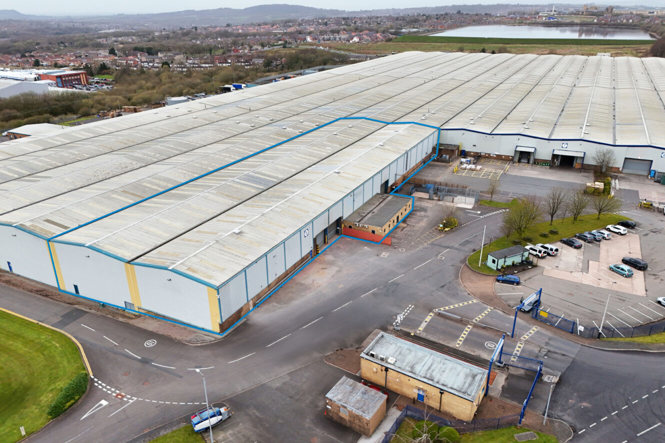 Aerial view of a large industrial warehouse complex with multiple sections, parking areas, and surrounding roads in a semi-urban area.