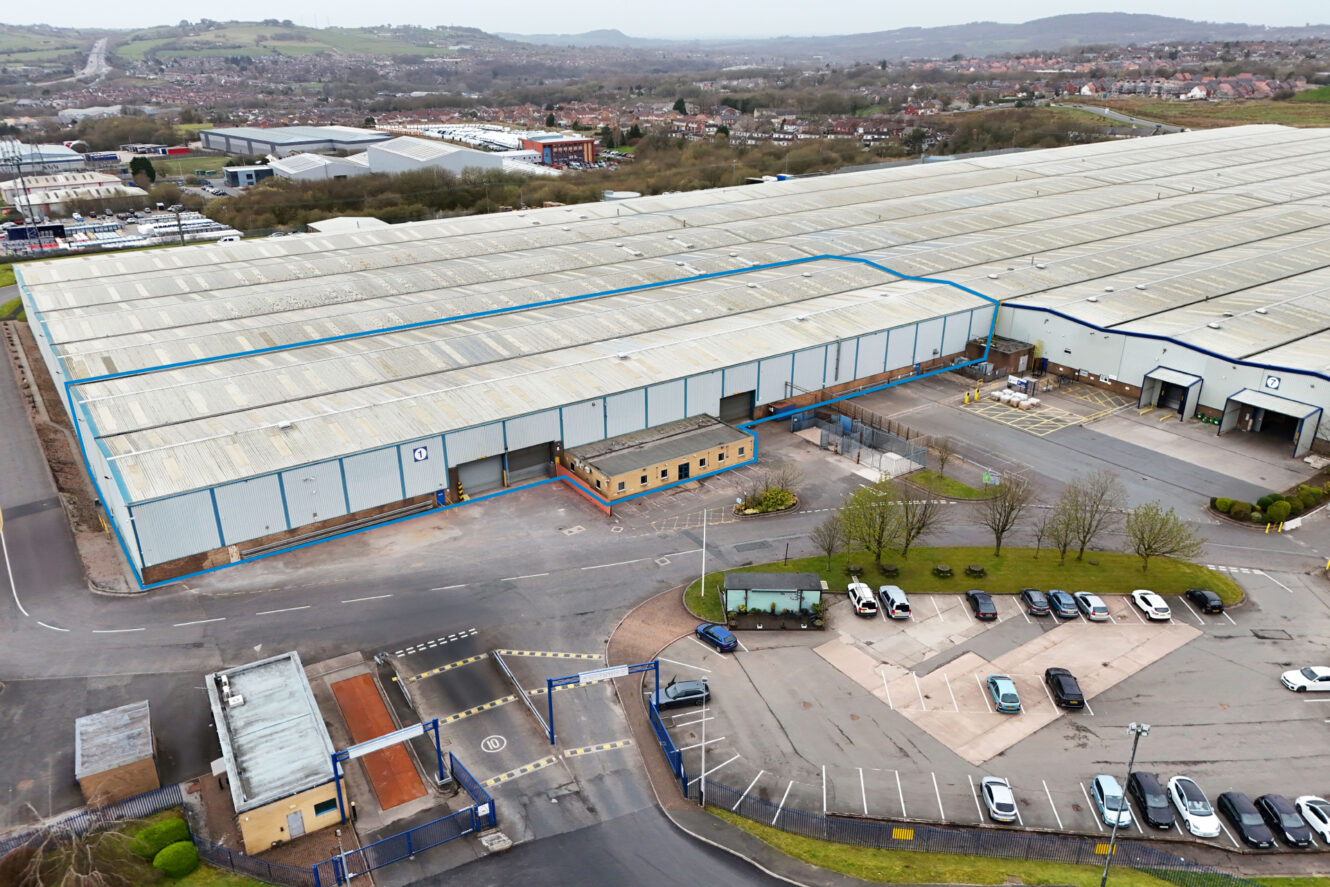 A large industrial warehouse complex with a marked section, a small office building in front, several parked cars, and surrounding roads and greenery.