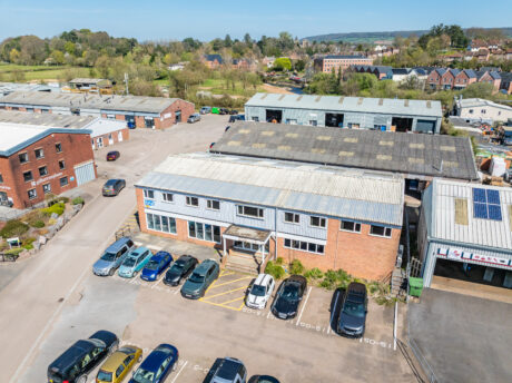Aerial view of an industrial estate with multiple warehouses, parked cars, and surrounding greenery under a clear sky.
