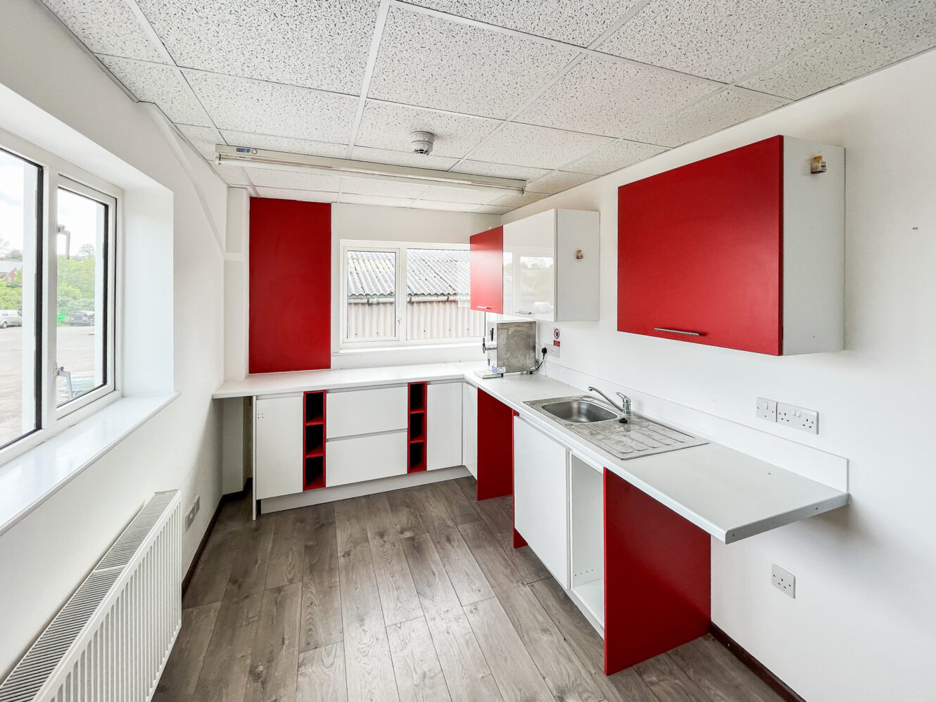 A small kitchen with white countertops, red and white cabinets, a stainless steel sink, and wood-look flooring, with windows letting in natural light.