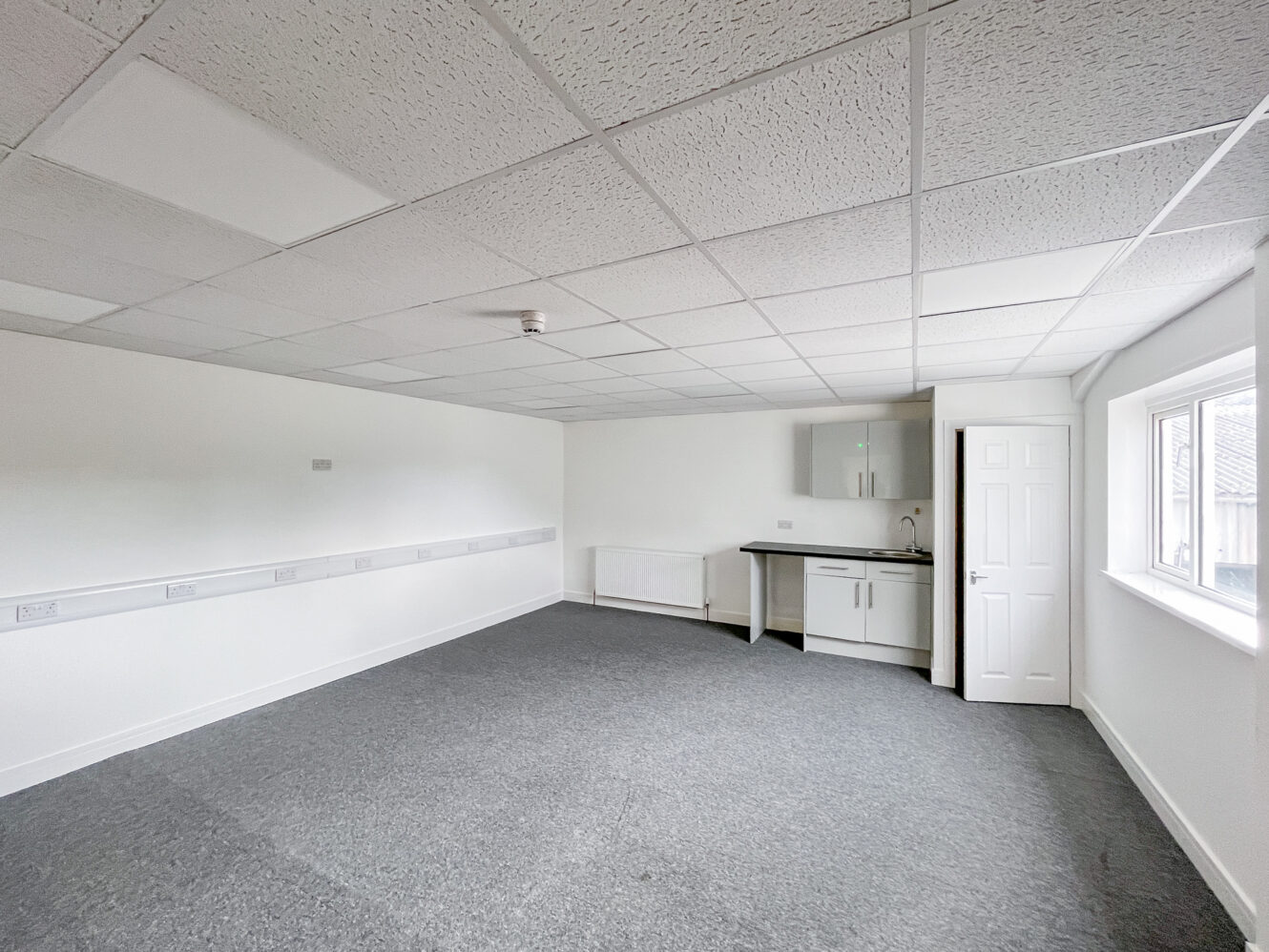 Empty office room with grey carpet, white walls, a ceiling with tiles, a small kitchenette, and a window letting in natural light.