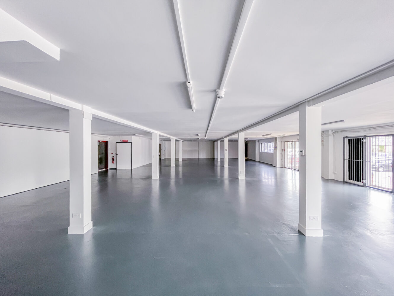 Large, empty industrial space with white walls, support columns, and gray concrete flooring, lit by overhead fluorescent lights and natural light from windows and glass doors.