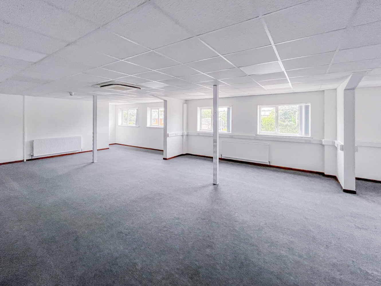 Empty office space with white walls, gray carpet, ceiling tiles, support columns, and large windows letting in natural light.