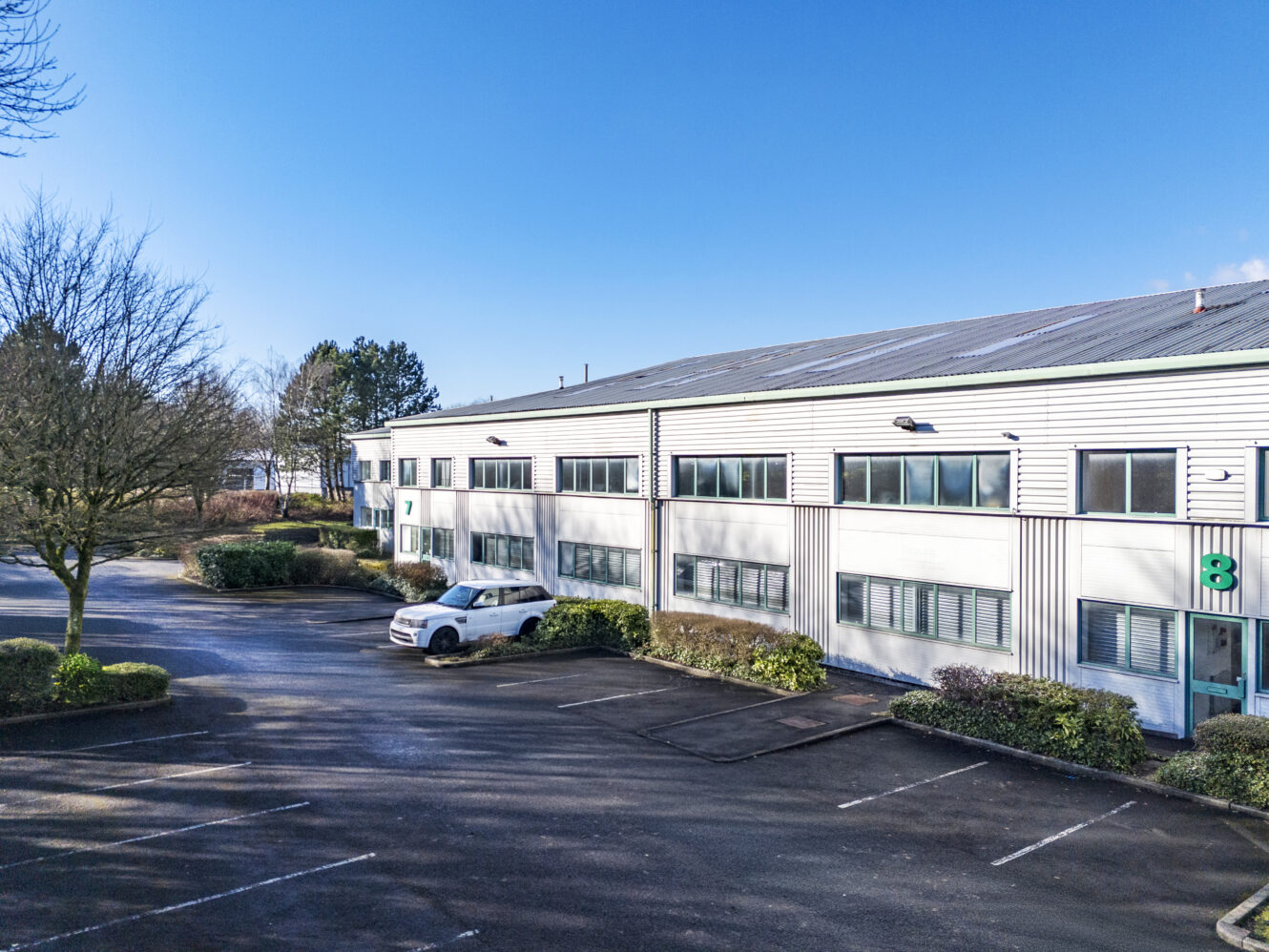 A two-story industrial or office building with numbered green doors, a white van parked outside, and an empty parking lot under a clear blue sky.