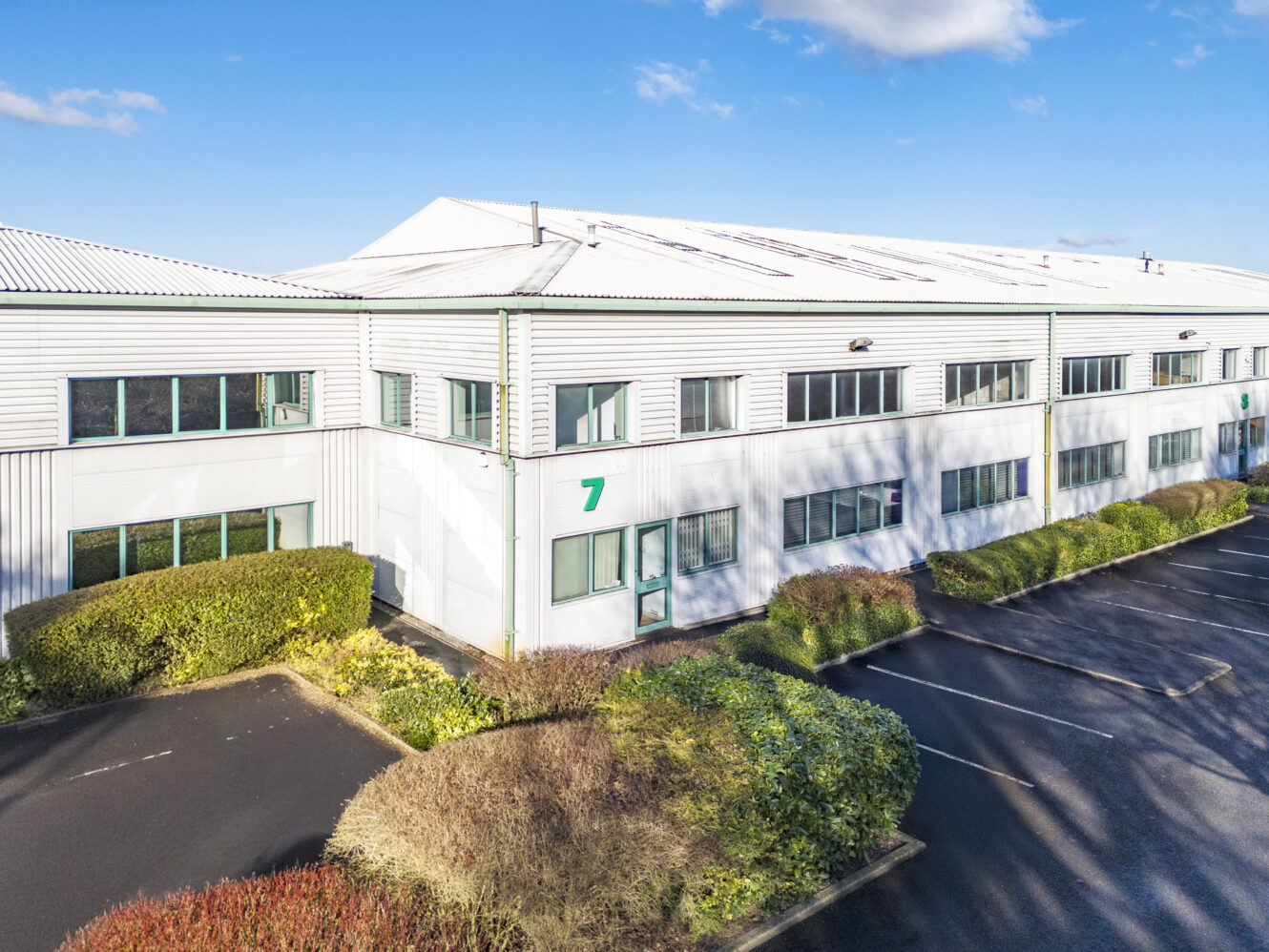 A large white industrial building with multiple windows, a green number 7 by the entrance, and bushes along the exterior under a clear blue sky.
