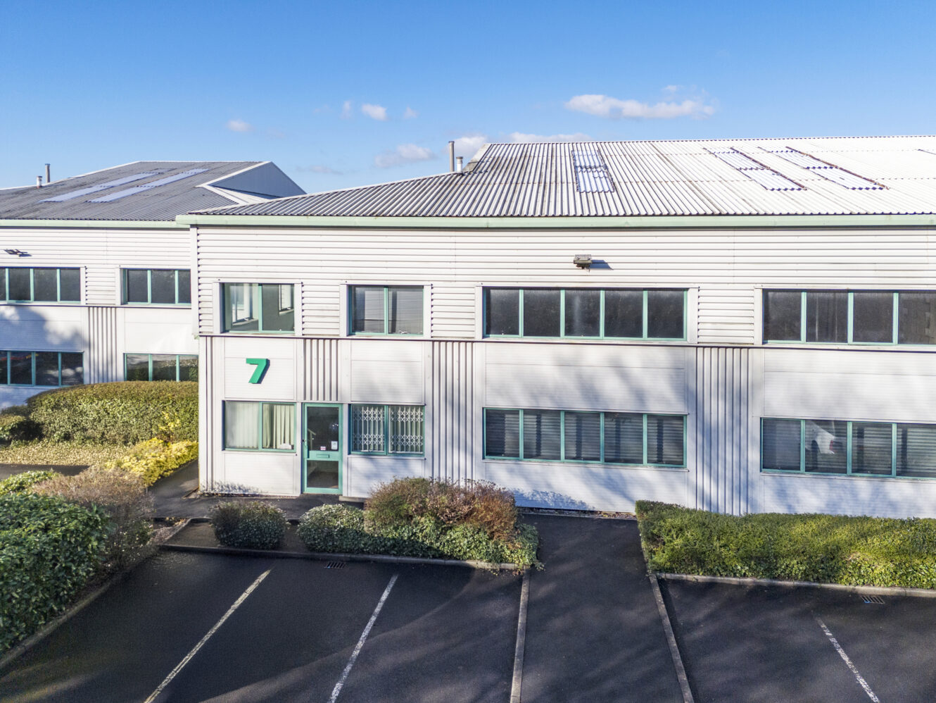 A modern industrial or office building with large windows, numbered 7, and surrounded by bushes and an empty parking area under a clear blue sky.