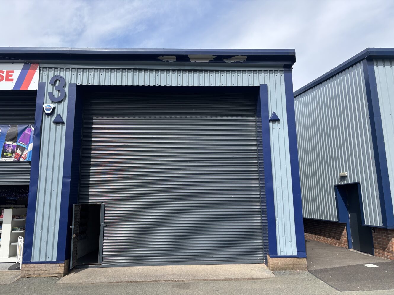A light blue industrial warehouse unit with a closed grey roller shutter door, labeled with number 3, and a small adjacent pedestrian door slightly open.
