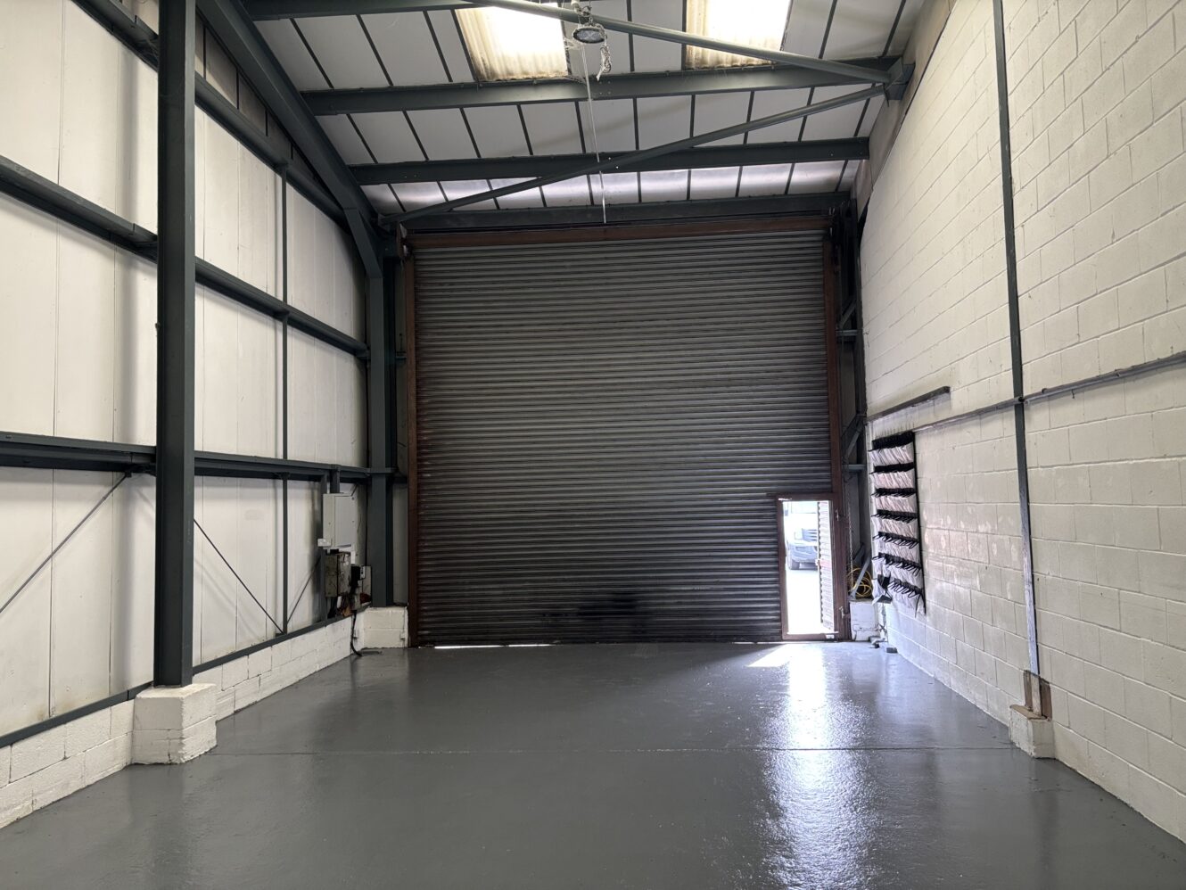 Empty industrial warehouse with gray painted floor, high ceiling, exposed beams, a large closed roller door, and a small open pedestrian door letting in daylight.