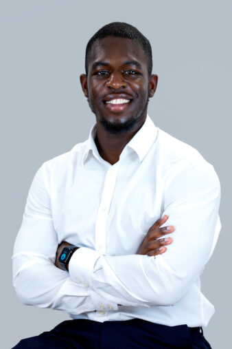 A man in a white dress shirt sits with arms crossed, smiling at the camera against a plain light gray background.