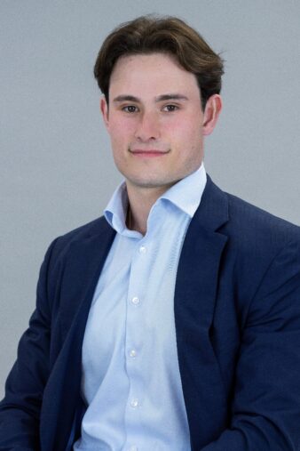 A young man with short brown hair wearing a navy blazer and light blue dress shirt sits facing the camera against a plain light background.