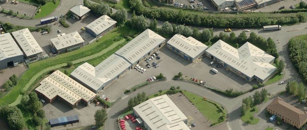Aerial view of an industrial area with several warehouse buildings, parking lots, surrounding greenery, and roads.