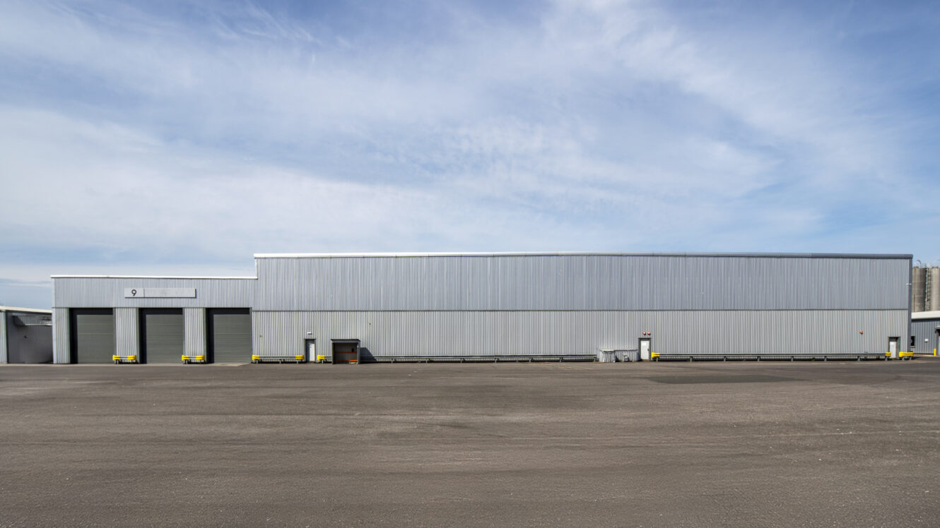 A large, gray industrial warehouse with multiple loading docks and a wide, empty asphalt area in front, under a partly cloudy sky.