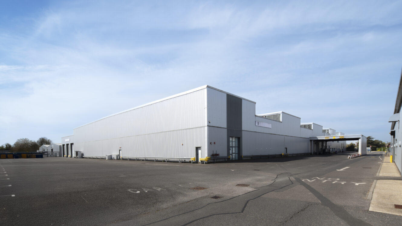 Large industrial warehouse with a gray exterior, surrounded by an empty paved lot under a clear sky.