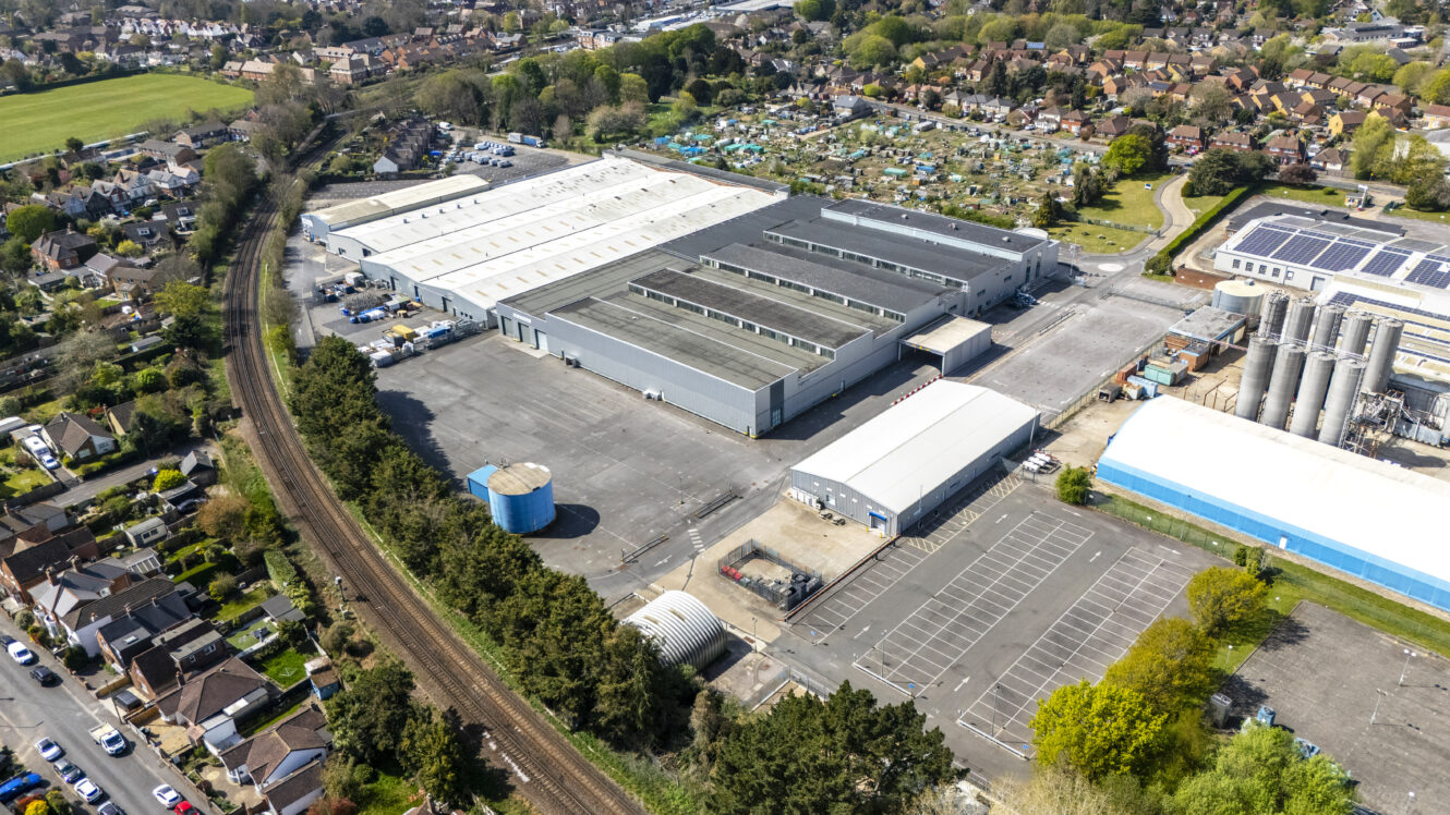 Aerial view of a large industrial facility with warehouses, parking lots, and nearby residential houses and railway tracks.