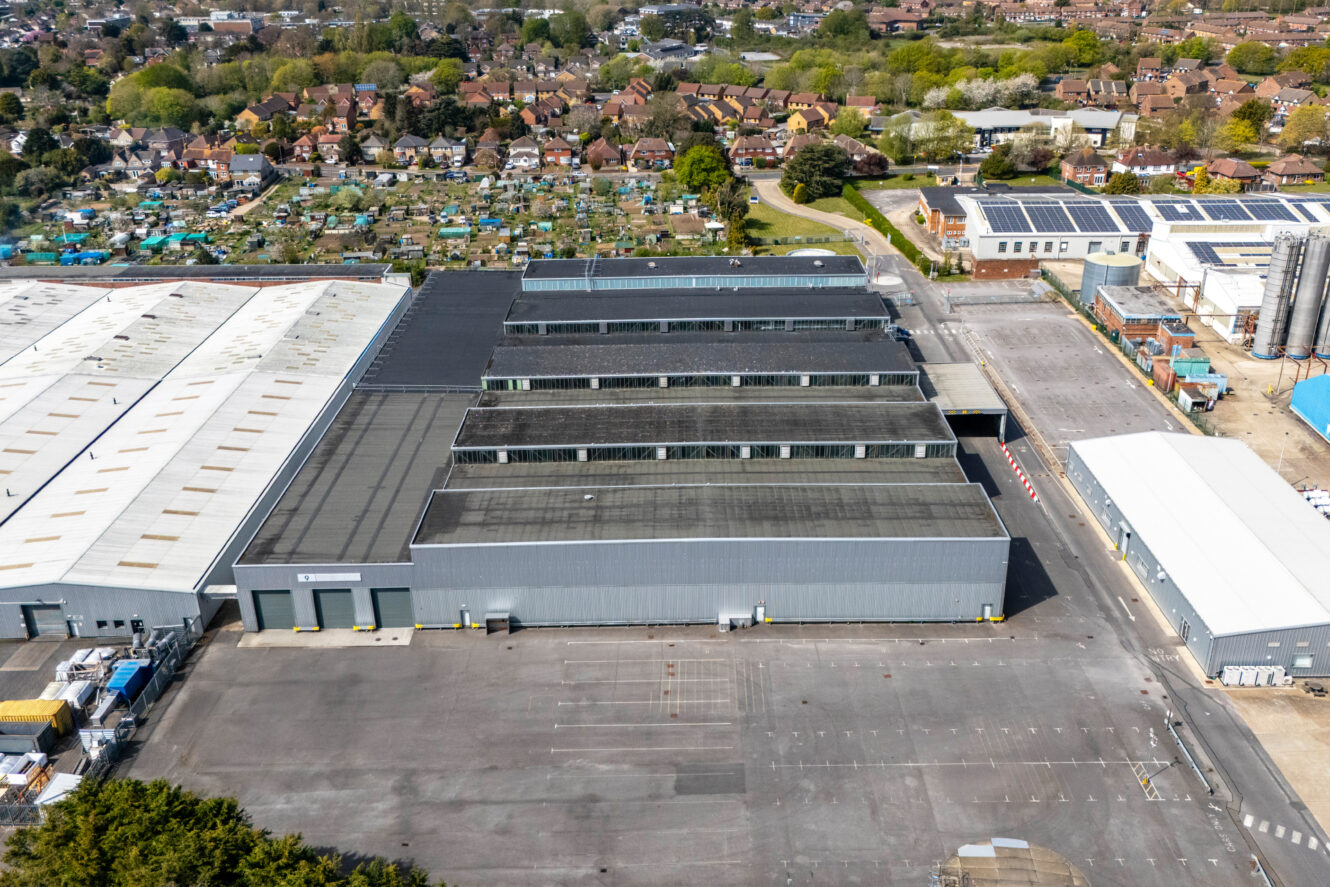 A large industrial warehouse with a dark roof and multiple loading bays, surrounded by empty paved areas and adjacent to other industrial buildings and a residential area in the background.