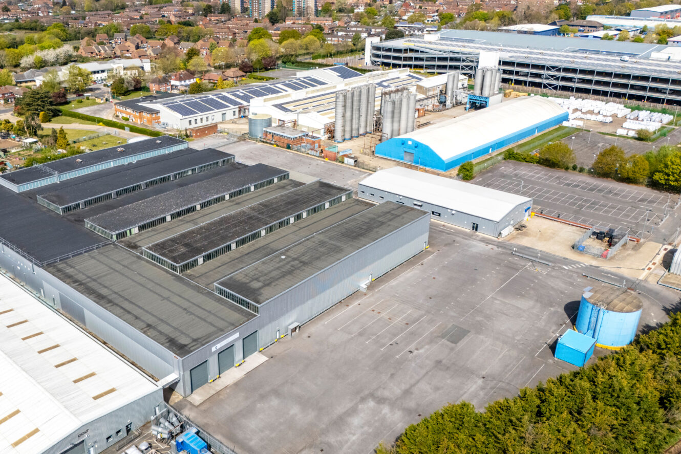Aerial view of an industrial complex with multiple large warehouse buildings, silos, a blue-roofed structure, and an adjacent parking lot, surrounded by trees and nearby residential areas.