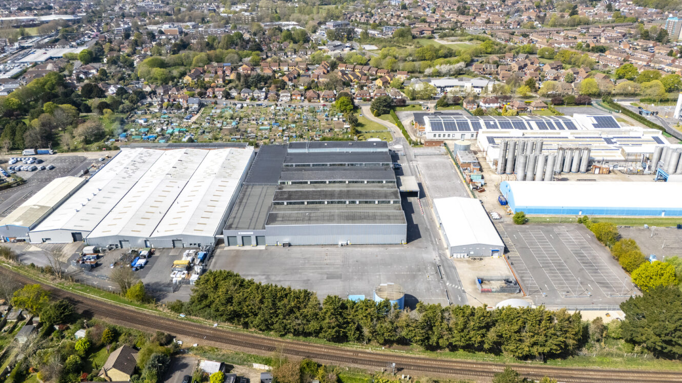 Aerial view of an industrial complex with large warehouses, parking lots, and adjacent railway tracks, surrounded by residential houses and green spaces.