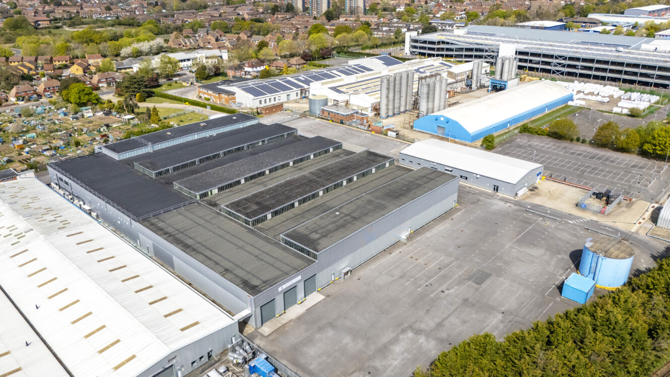Aerial view of an industrial complex with multiple large warehouses, storage facilities, parking areas, and nearby residential housing.