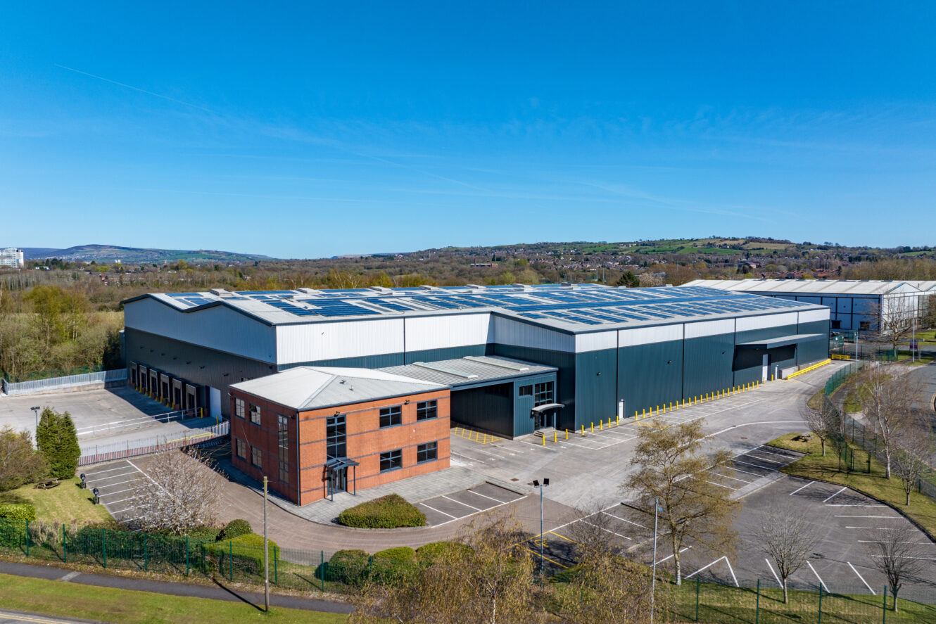 A large industrial warehouse with a brick office building in front and an adjacent parking lot, set on a clear day with blue sky and distant hills.