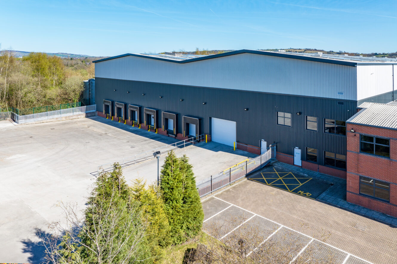 A large industrial warehouse with multiple loading docks, an empty parking area, and surrounding trees under a clear sky.