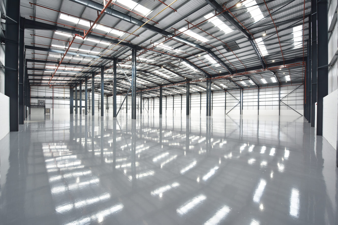 Empty modern warehouse with high ceilings, polished concrete floors, exposed steel beams, and large windows allowing natural light inside.