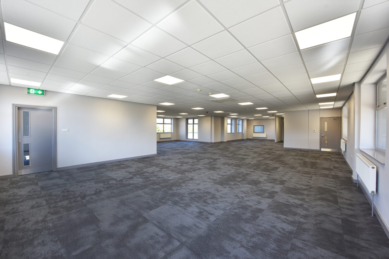Large empty office space with gray carpet, white ceiling tiles, fluorescent lights, and multiple windows along the walls.