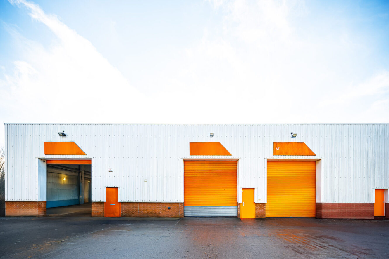 A white industrial warehouse with orange doors and accents, featuring three large garage doors and two entry doors, on a paved lot under a clear sky.