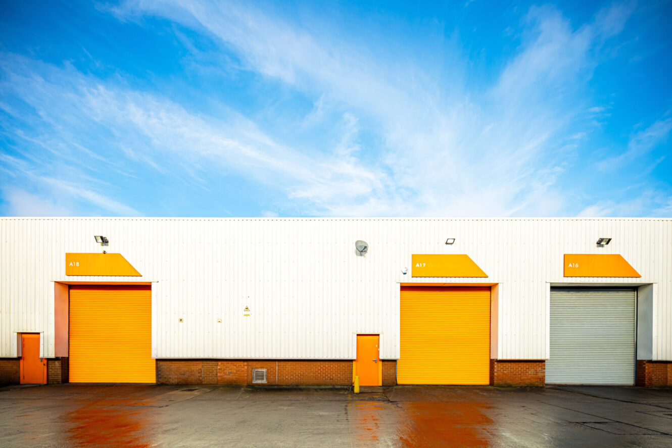 A white industrial warehouse with three large doors—two orange and one gray—under a blue sky with wispy clouds.