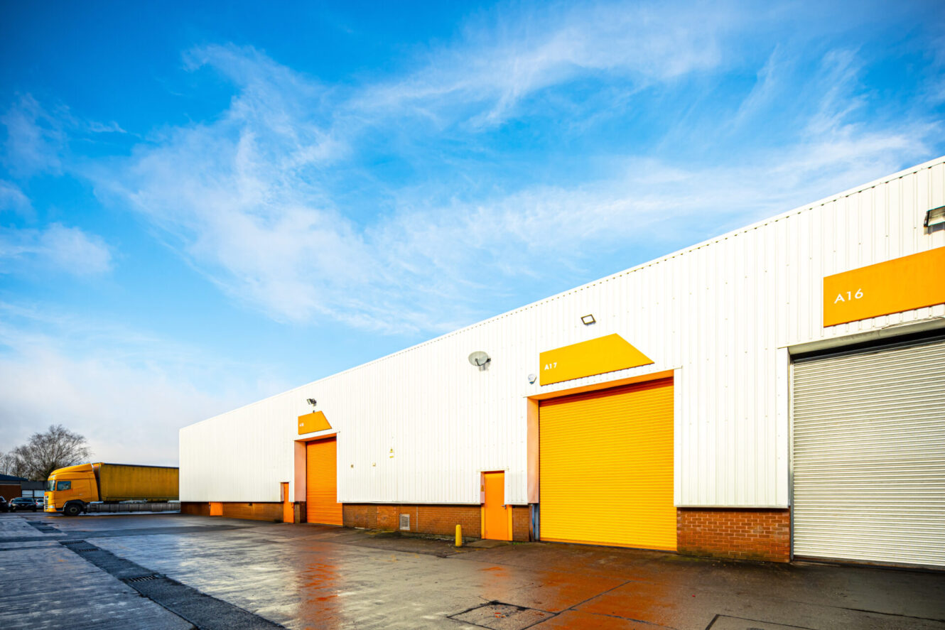 Row of industrial warehouse units with yellow and white exteriors, each featuring large yellow roller doors, on a paved lot under a partly cloudy sky.
