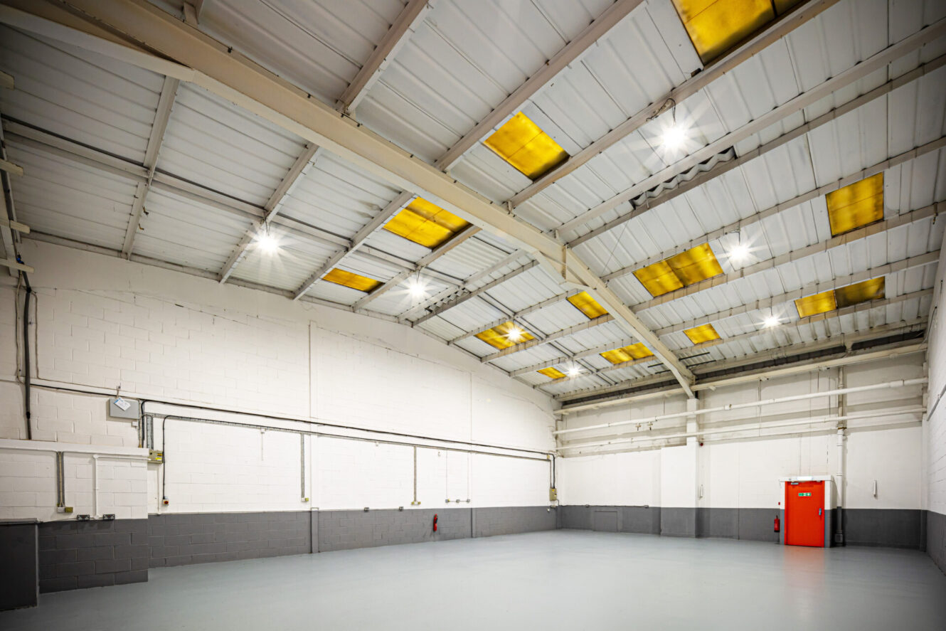 Empty industrial warehouse interior with white and gray walls, exposed ceiling beams, rows of overhead lights, and a single bright orange door on the right side.