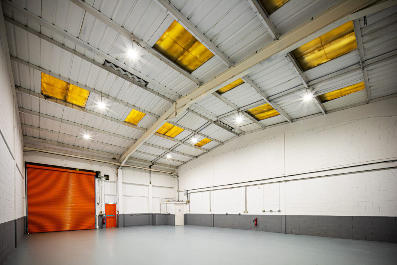 Empty industrial warehouse with a grey floor, white and grey walls, orange shutter door, and yellow-tinted skylights in the ceiling. Bright overhead lighting is on.