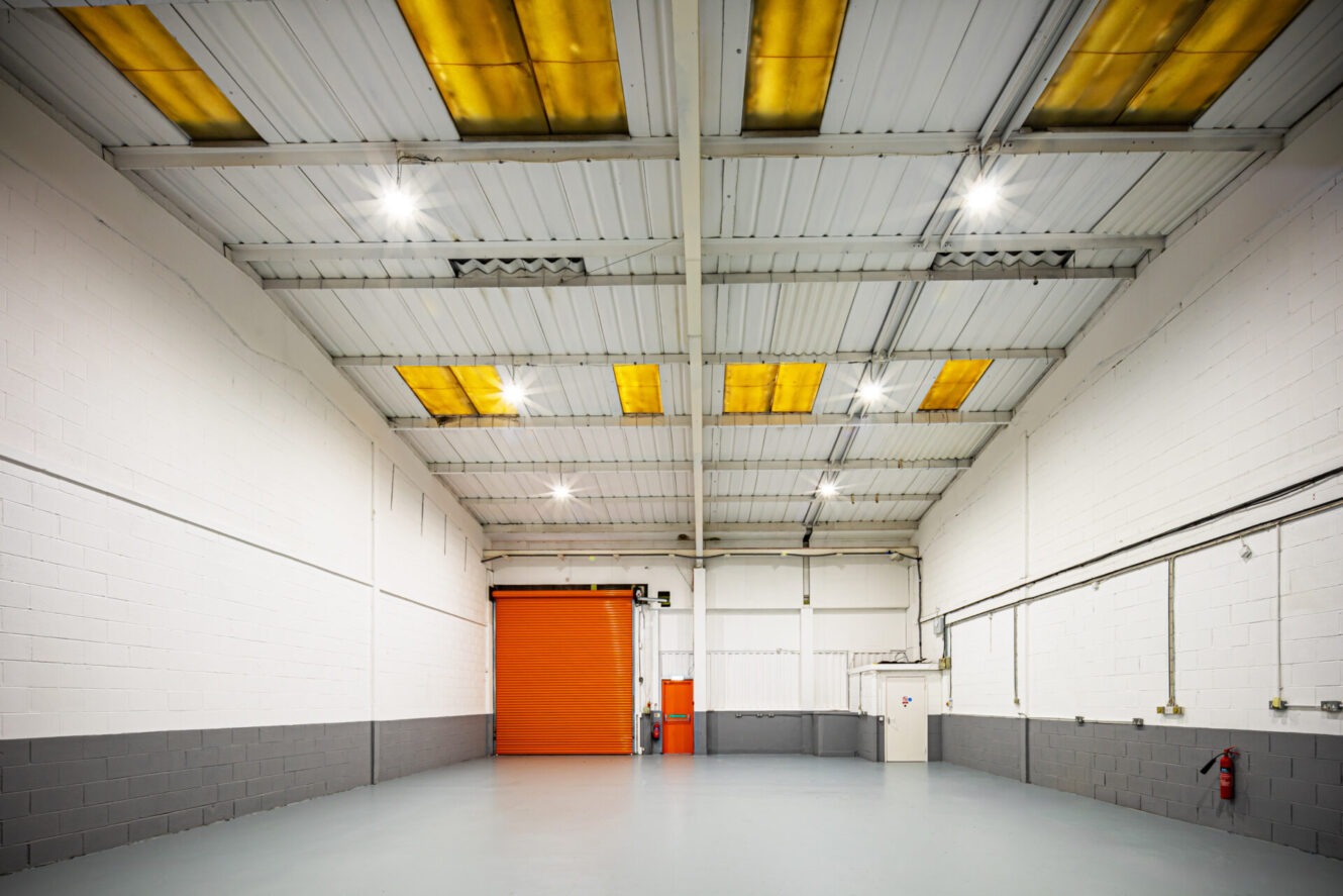 Empty industrial warehouse interior with white and gray walls, orange roller shutter door, exposed ceiling beams, and fluorescent overhead lighting.