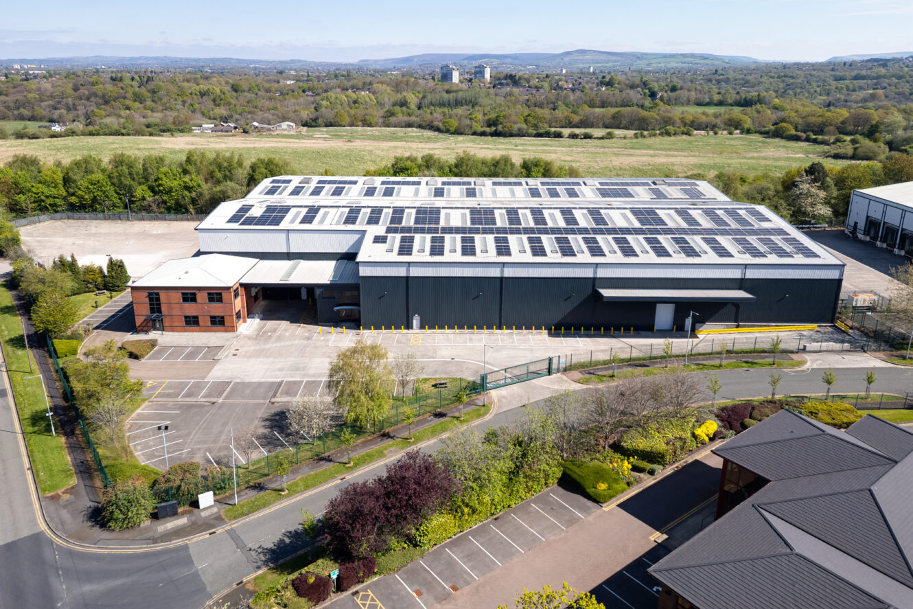 A large industrial warehouse with solar panels on the roof, surrounded by parking lots and greenery, with hills visible in the background.