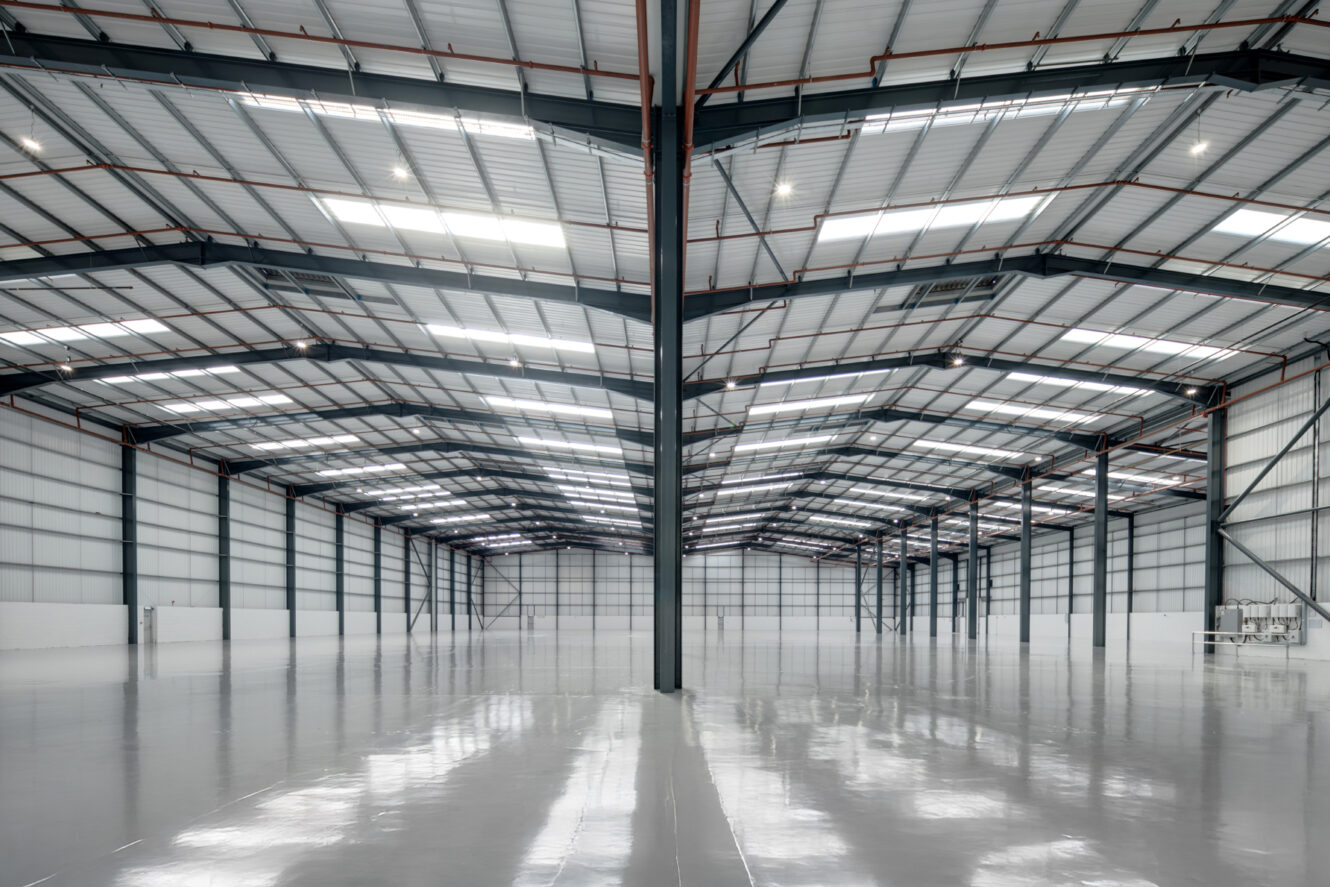 Interior of a large, empty warehouse with polished concrete floors, high metal ceilings, and evenly spaced support beams under bright lighting.