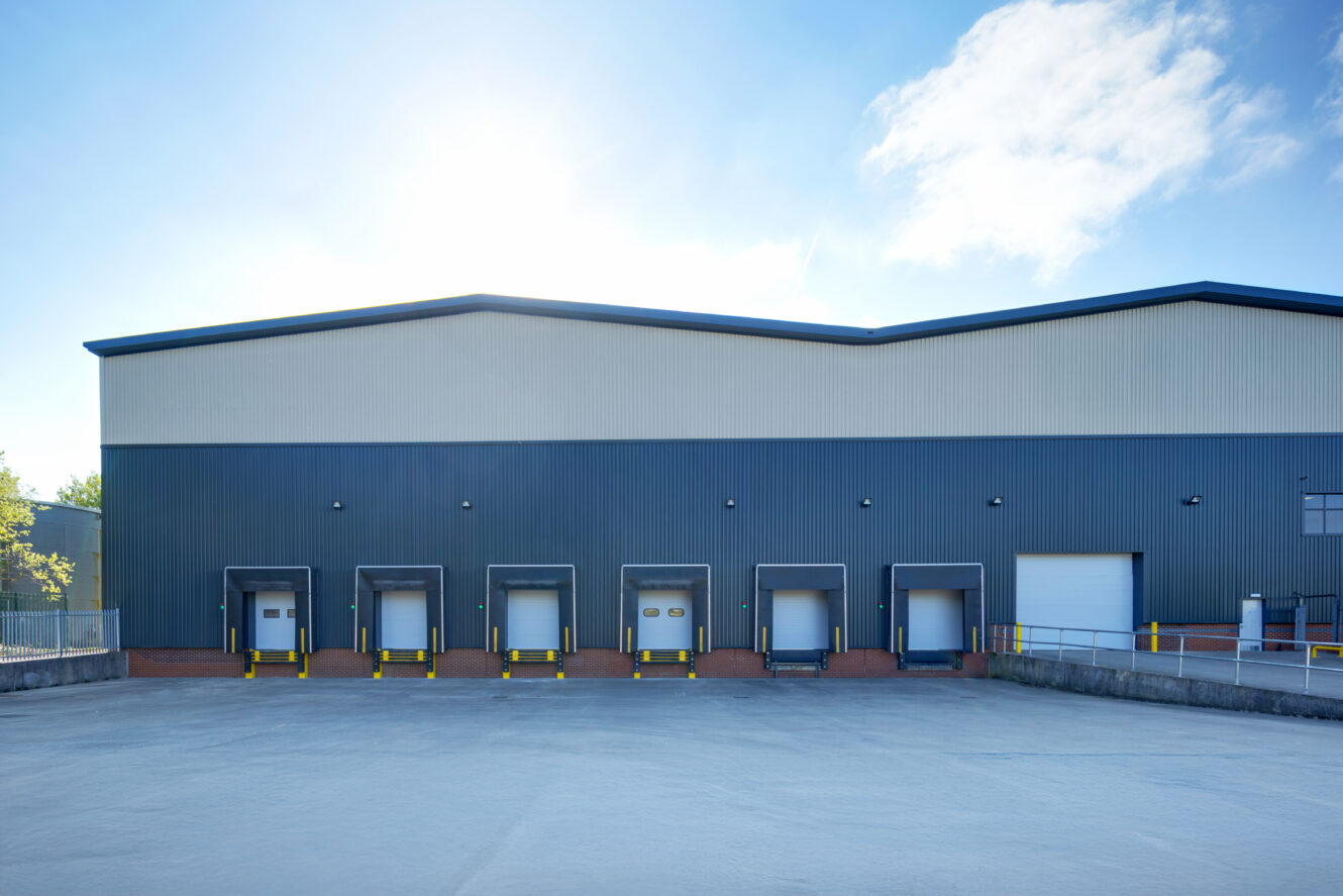 A warehouse exterior with five loading dock doors and one large white door, situated in a paved lot under a clear sky.
