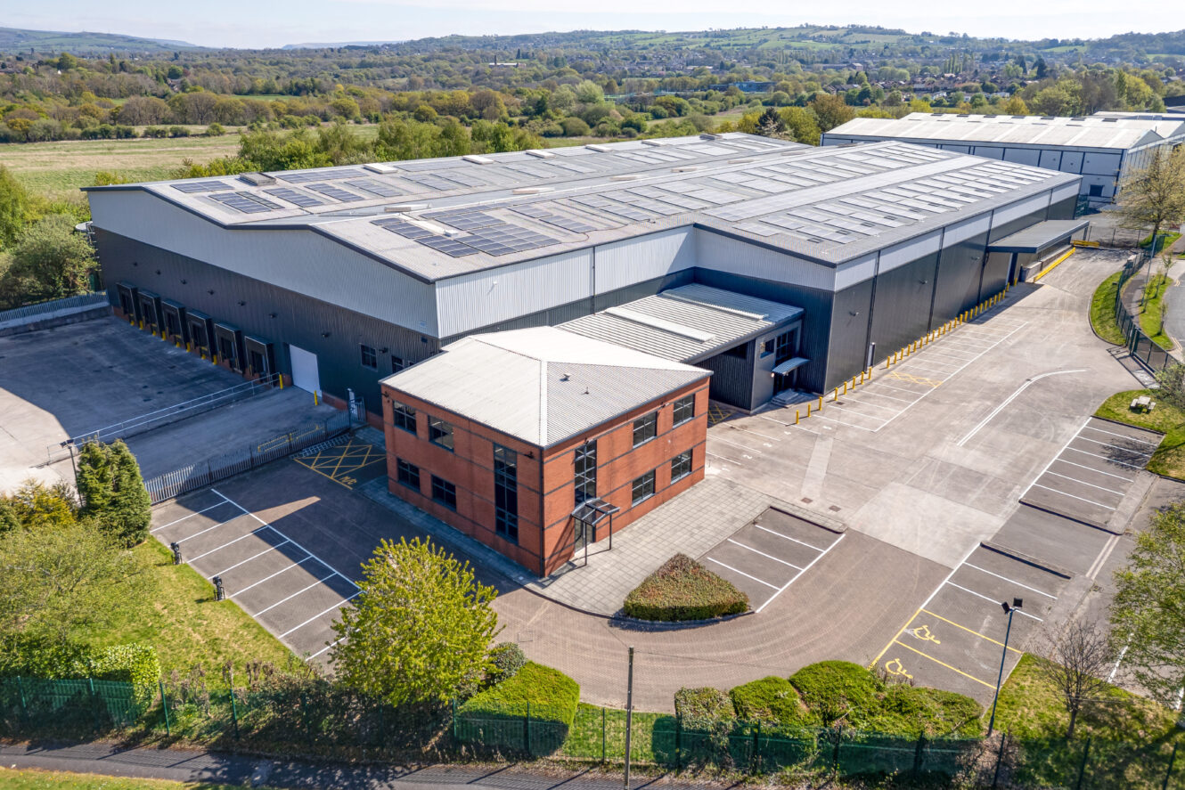 A large warehouse with solar panels on the roof, adjacent to a smaller brick office building, surrounded by parking spaces and greenery.