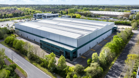 Aerial view of a large, modern industrial warehouse with a parking lot, surrounded by trees and nearby buildings.