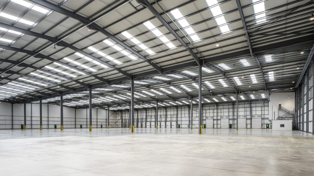 Spacious, empty warehouse interior with high ceilings, exposed steel beams, skylights, concrete floors, and large industrial doors.