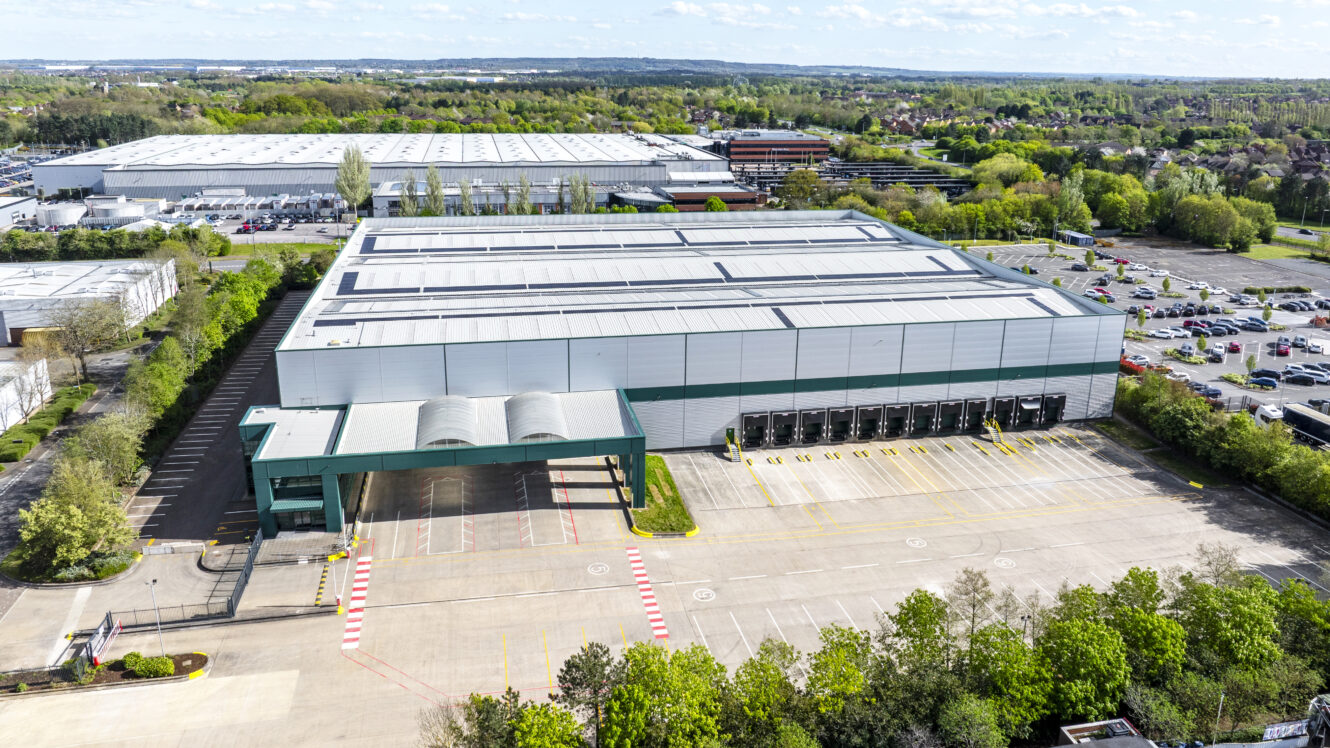 Aerial view of a large warehouse facility with multiple loading docks, adjacent parking areas, and surrounding industrial buildings and greenery.