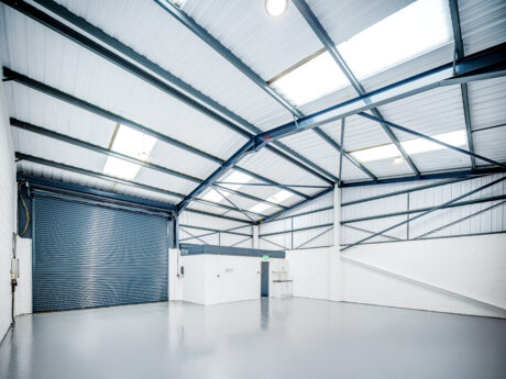 Empty industrial warehouse with white walls, metal ceiling beams, skylights, polished gray floor, and a large closed roller door on one side.
