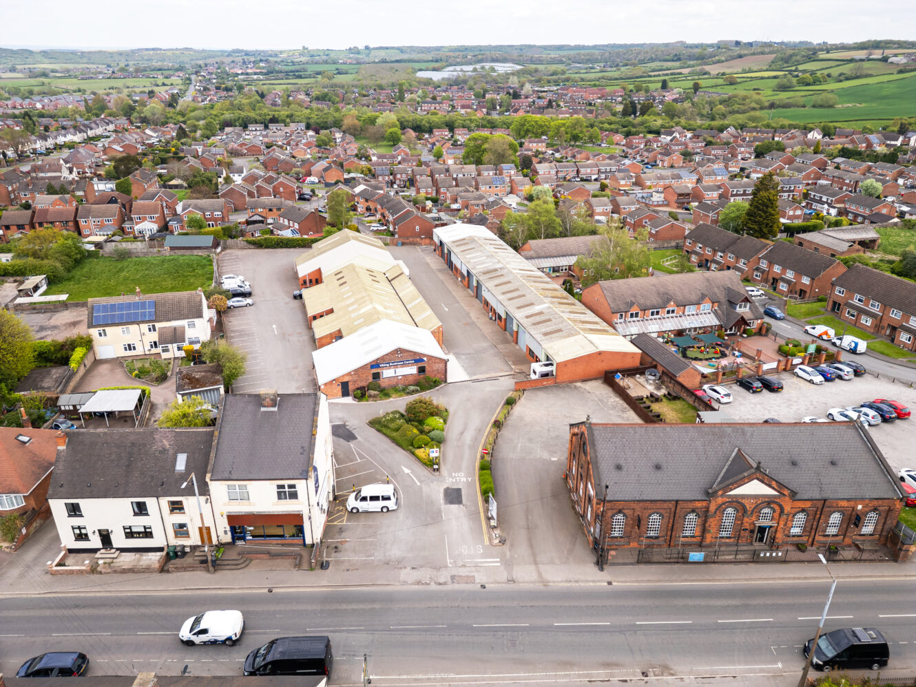 Aerial view of a small town with residential houses, industrial buildings, parked cars, and green fields in the background.