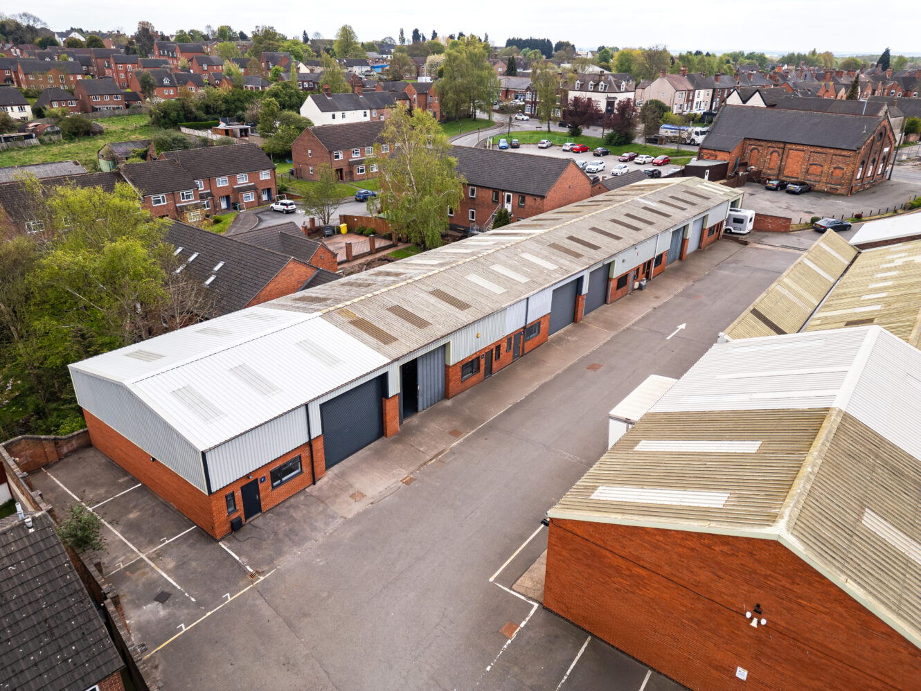 Aerial view of a row of industrial warehouse units with roller doors, surrounded by parking spaces, set in a suburban area with houses and trees.