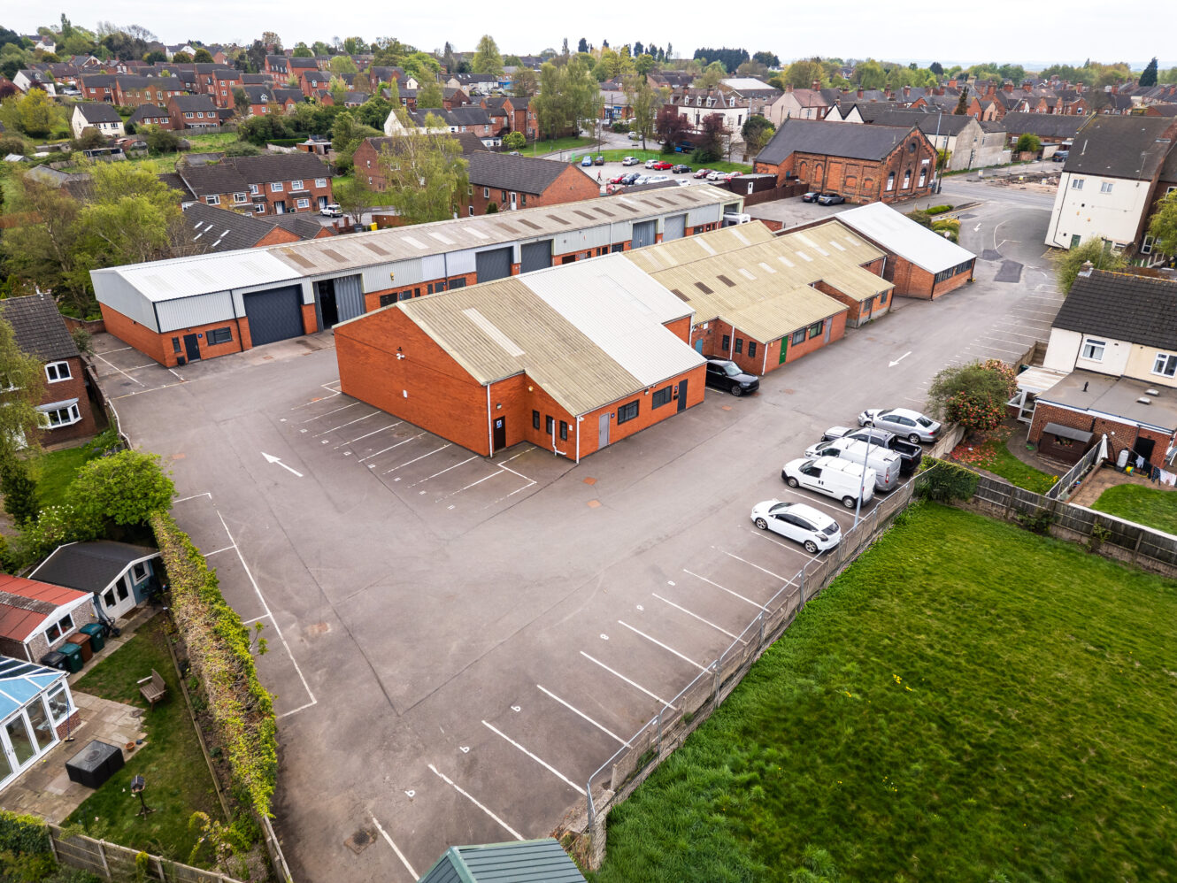 Aerial view of a small industrial complex with several warehouse buildings, a parking lot with parked cars, and surrounding residential houses and green spaces.