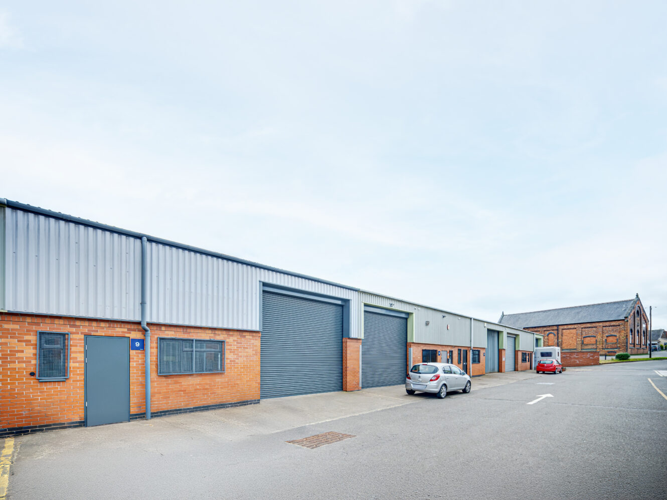 A row of industrial warehouse units with brick and metal exteriors, large roller doors, and two parked cars on an asphalt lot.